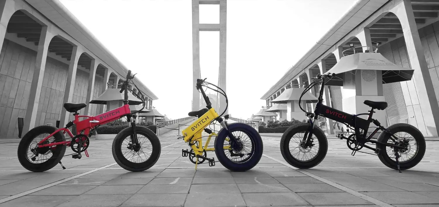 Three Svitch electric bicycles in red, yellow, and black, displayed outdoors against a modern architectural backdrop